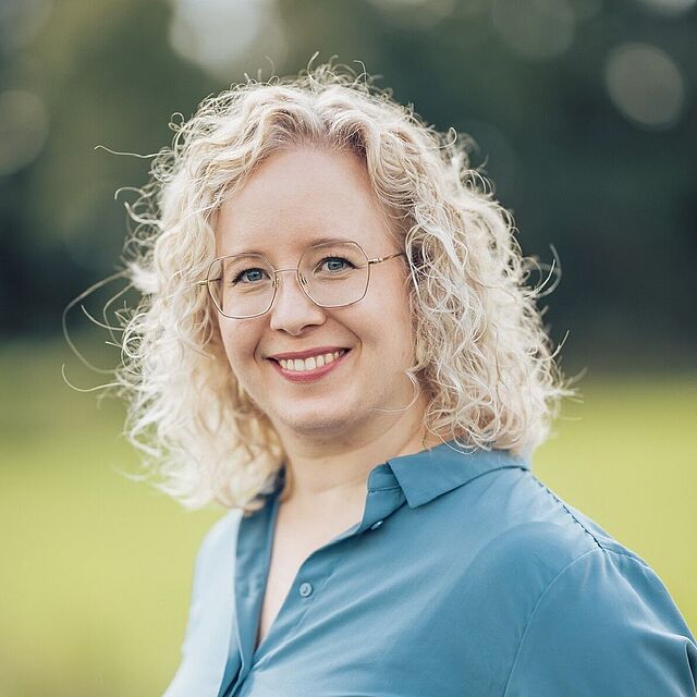 A woman with curly blond hair and glasses is smiling outdoors